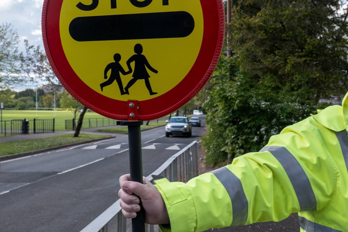 An individual in a reflective safety jacket is holding a circular road sign with the word "STOP" written in white capital letters on a red background. The sign also features a yellow circle with a depiction of two children walking, intended to indicate a pedestrian crossing. The scene is set by a road with visible traffic and greenery in the background.