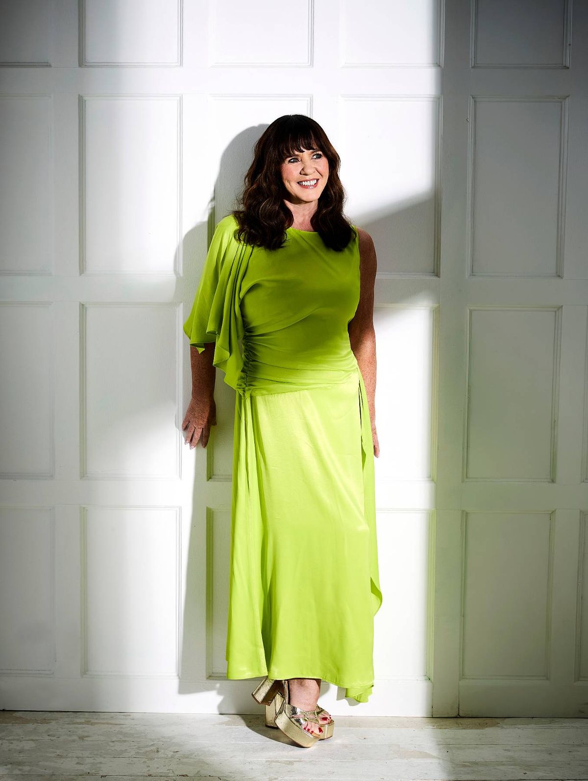 An individual, dressed in a vibrant lime green garment, stands against a backdrop of a white, paneled wall. The interplay of light and shadow casts a distinctive silhouette against the wall, highlighting the subject's presence in the scene.