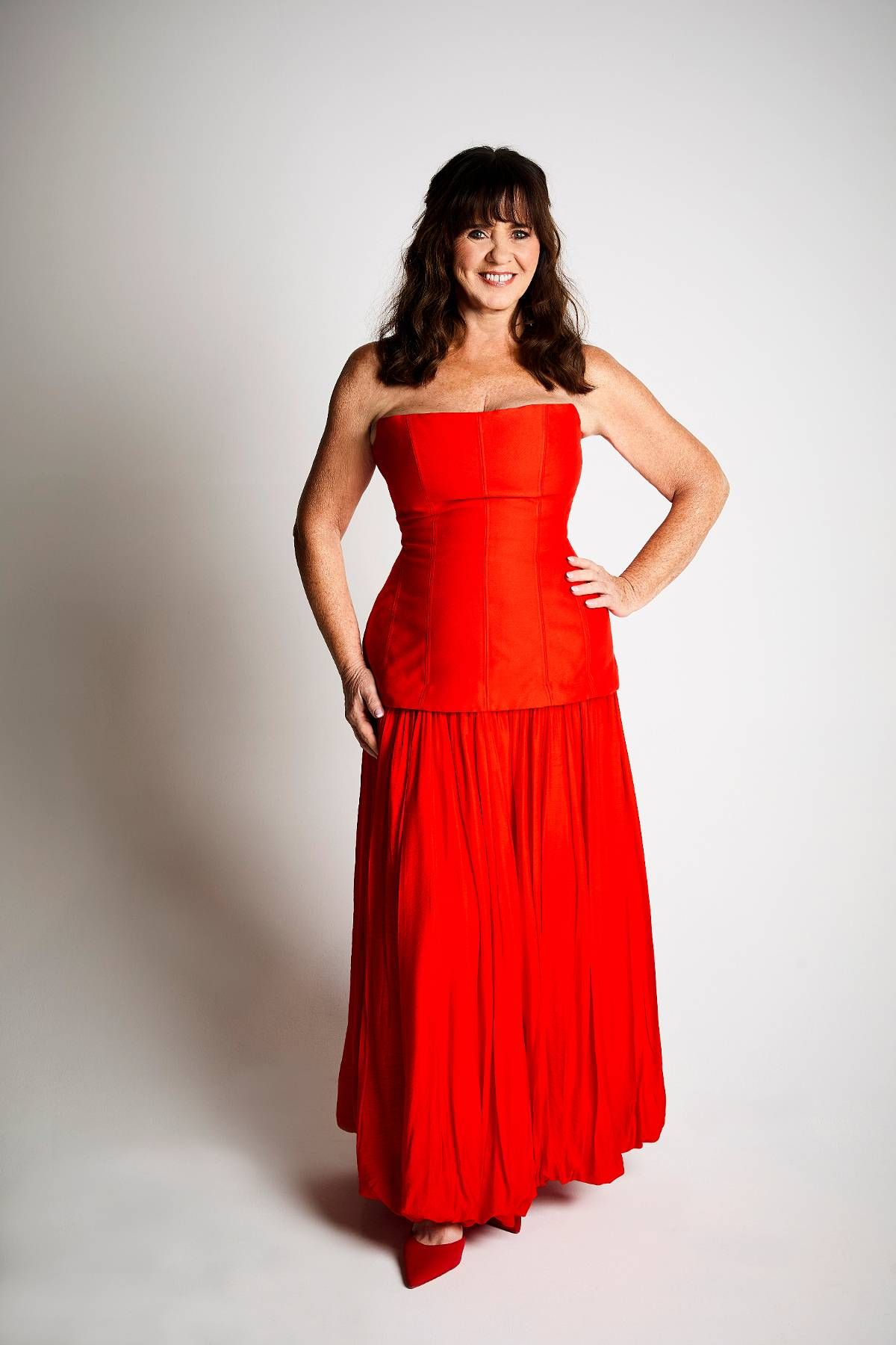 An individual is depicted in a portrait, standing with one hand on their hip and a slight smile. They are dressed in a vibrant red strapless dress. The background is plain and white, with minimal visual distractions.