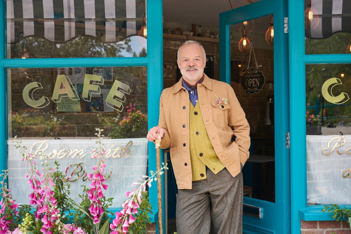 An elderly gentleman is standing in front of a vibrant blue entrance to a café, dressed in a tan jacket and gray trousers. The café’s exterior is adorned with colorful flowers and has a glass door with the word "CAFE" prominently displayed in illuminated letters.