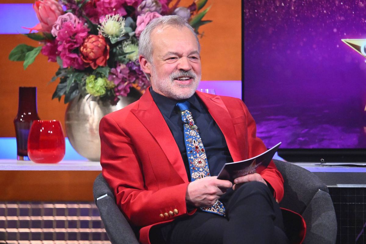 An individual dressed in a red blazer, black tie, and black trousers is seated in a chair, holding a book and smiling. Behind them, there is a large television screen displaying a vibrant background, and a floral arrangement is visible on a nearby table.
