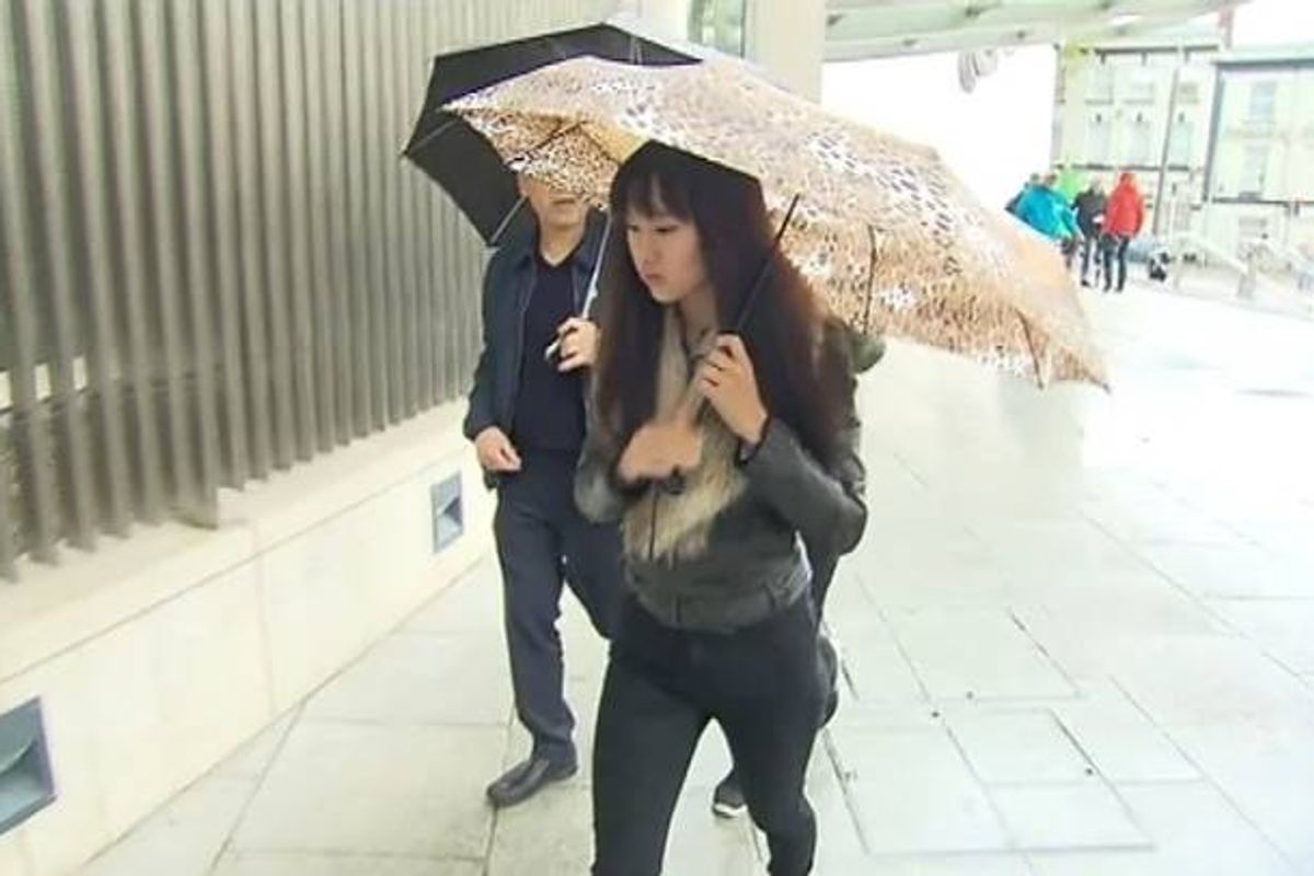 An individual in a black jacket holds an umbrella, standing beside another person under the rain, with a building and pedestrians in the background.