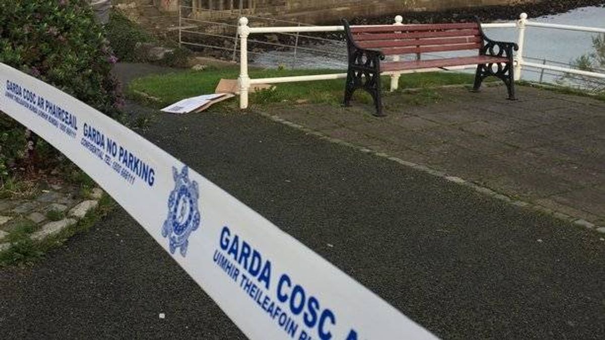 An image depicts a paved area cordoned off with a white ribbon bearing the words "Garda COSC" and a logo. In the background, a bench and a white railing are visible, with a grassy area and a body of water beyond.