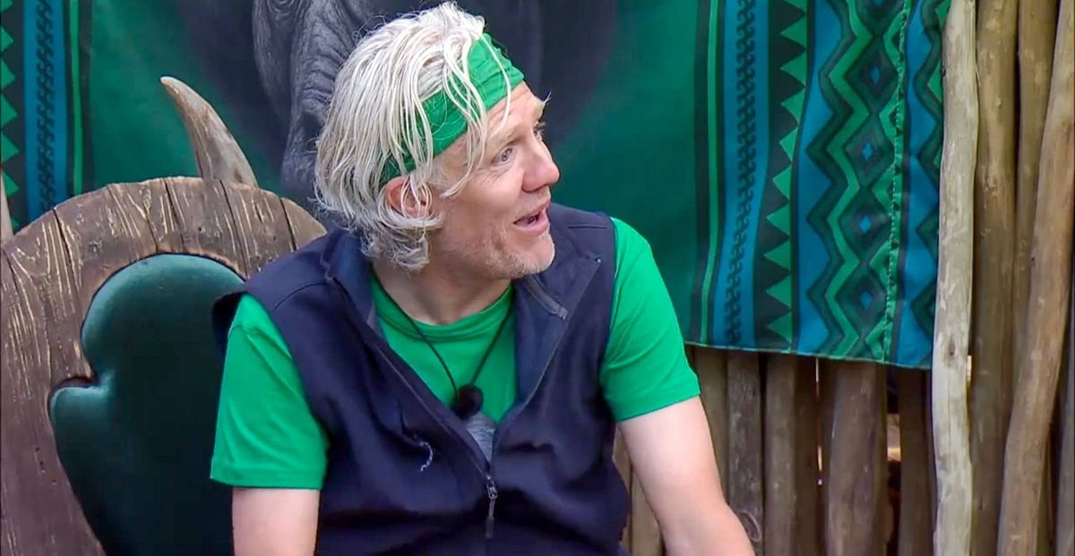 An older individual with white hair and a green headband is seated in a chair against a backdrop adorned with green foliage and a large, intricate wooden design. They are dressed in a green shirt and a dark blue vest, and appear to be engaged in conversation.
