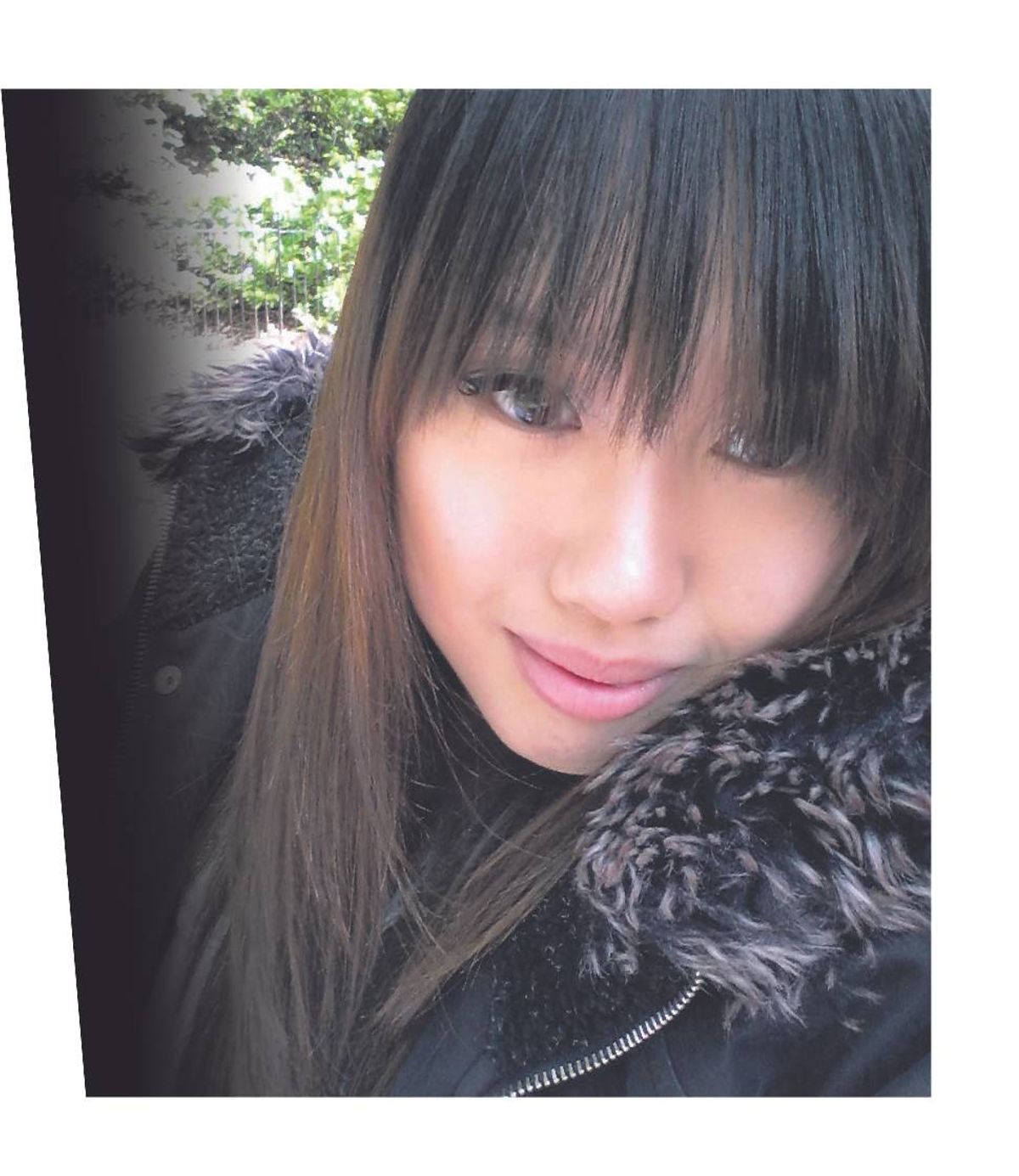 A close-up portrait of an individual with long, straight black hair and bangs, captured from a low angle. The person is wearing a dark-colored, fur-trimmed jacket and is smiling gently. The background is blurred with natural elements.