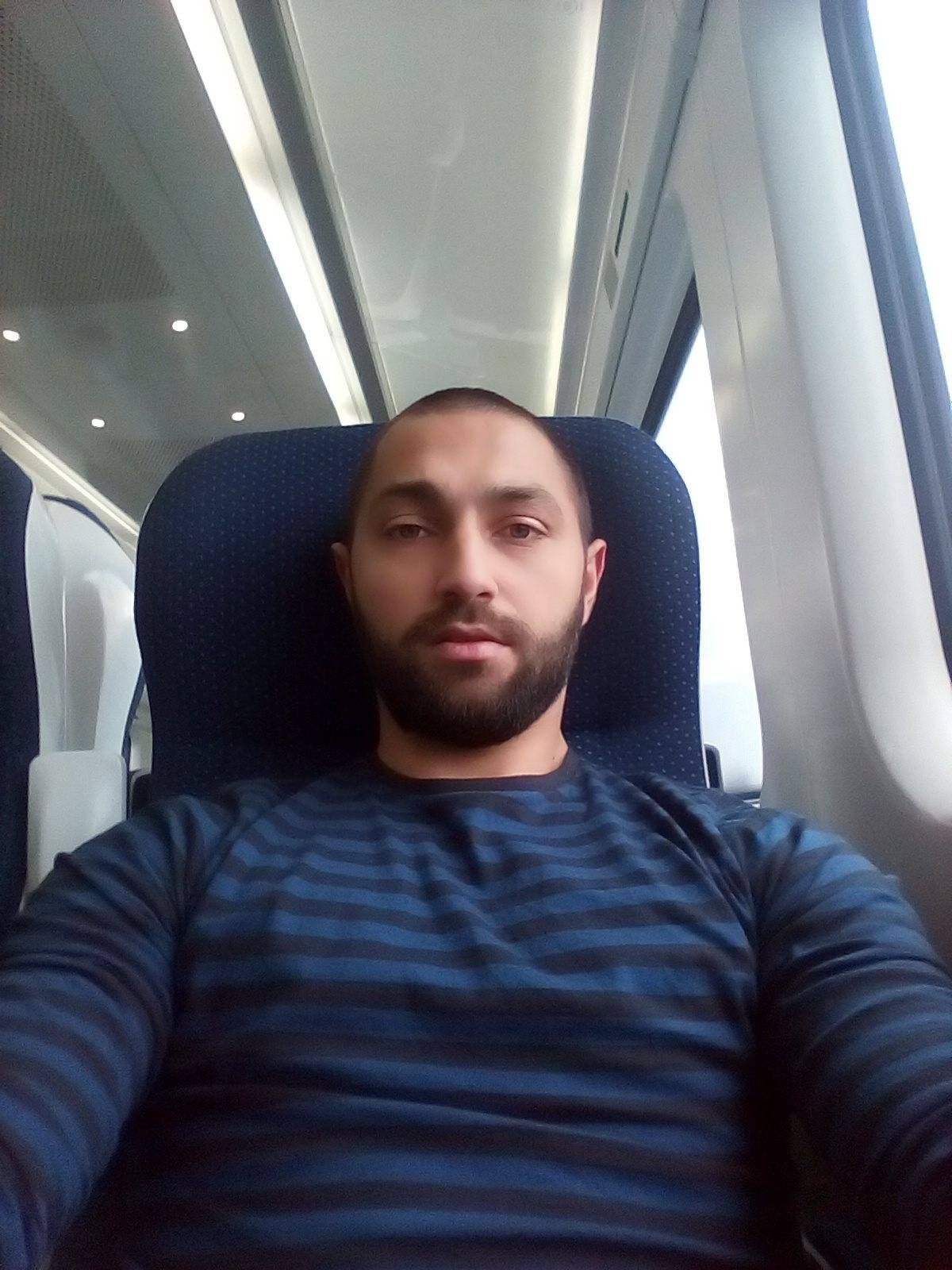 An individual is seated on a blue chair inside a vehicle, possibly a train, appearing to be asleep. The setting is illuminated with ambient lighting, and the individual is wearing a dark blue shirt.