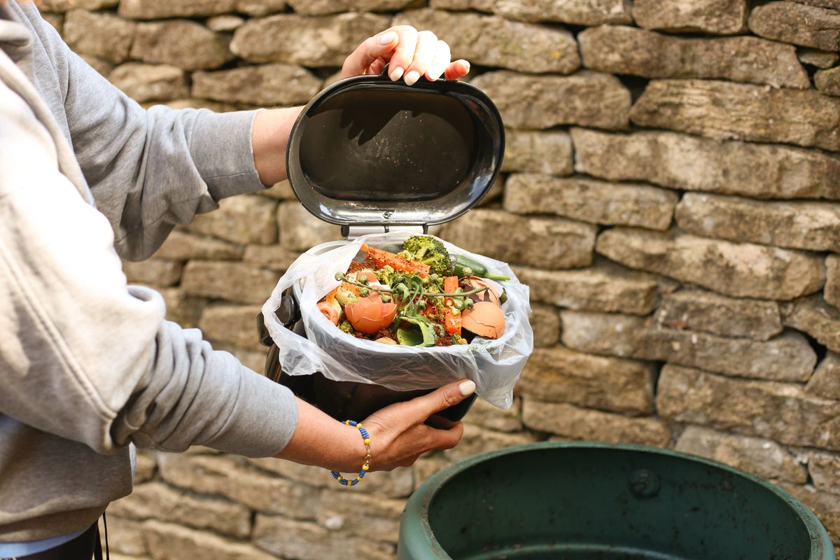 A person holds a container full of kitchen scraps—vegetable peels, eggshells, and food leftovers—above a green compost bin outdoors. This image captures a practical and eco-friendly moment in the composting process, promoting sustainable living and reducing food waste.