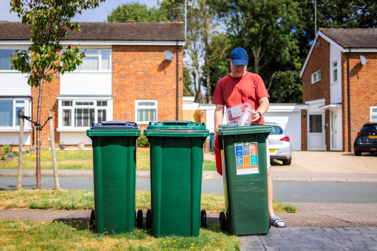 Adult male putting plastic waste into his recycling bin on a residential street, awaiting collection. It is summer and the man is dressed casually in beige shorts and red t shirt.