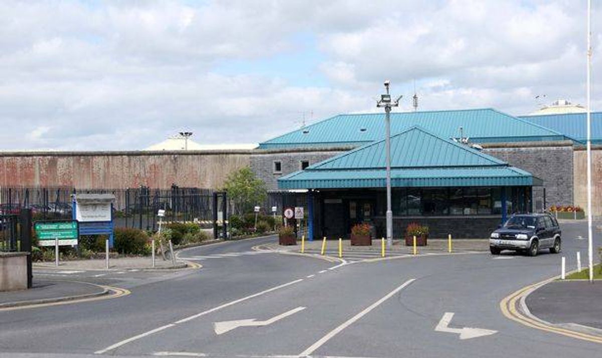 An image depicts a road leading to a building with a blue roof. The road is empty, and there is a car parked at the side. The area is surrounded by a fence, and there are several plants and a pole with a flag. The sky is cloudy.