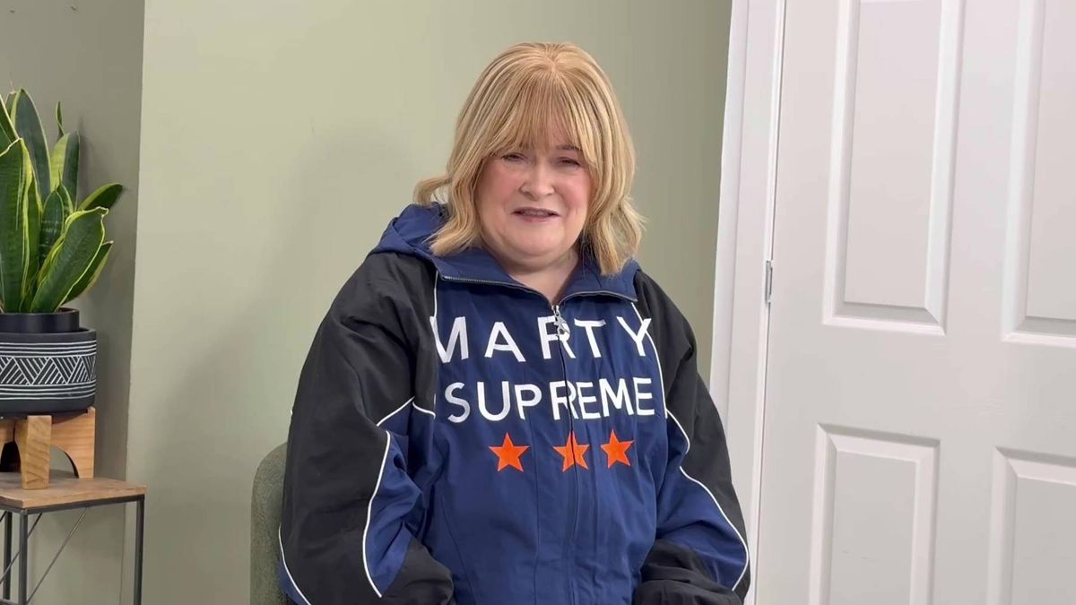An individual with blonde hair is seated and wearing a blue and black hoodie jacket adorned with the text "MARTY SUPREME" and red stars. The person is positioned in front of a white door and a small table with a potted plant.