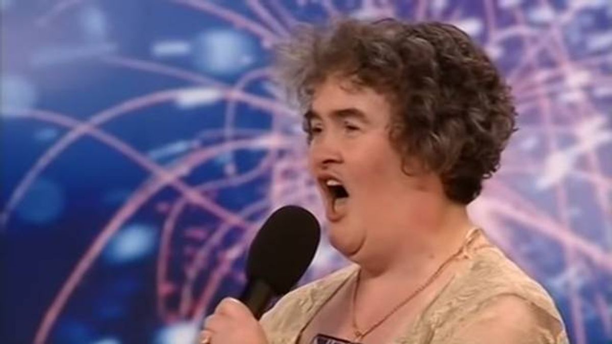 An individual, with short curly hair, is speaking into a microphone. The person is dressed in a light-colored top and has a necklace. The background features a vibrant array of colors and intricate patterns, suggesting a lively or artistic setting.