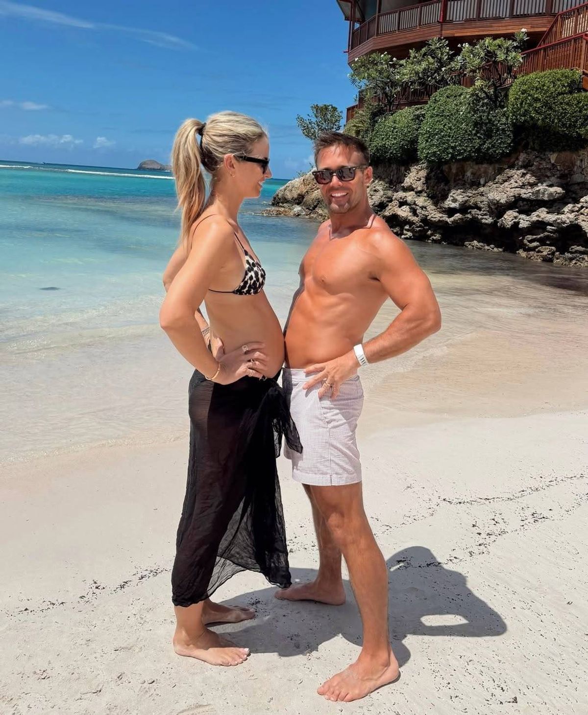 A man and a woman are standing on a sandy beach, posing for a photograph. They are both dressed in summer attire, with the woman wearing a bikini and the man shirtless. Behind them, there is a scenic backdrop featuring a rocky shoreline and lush greenery.