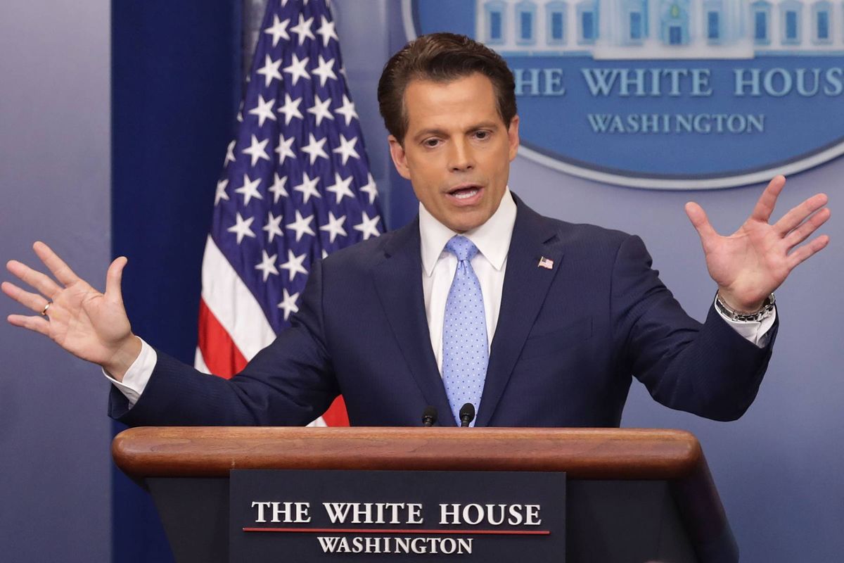 An individual dressed in a formal suit, standing at a podium, gestures with his hands while addressing a crowd. The podium is labeled "The White House" and an American flag is positioned behind the speaker.