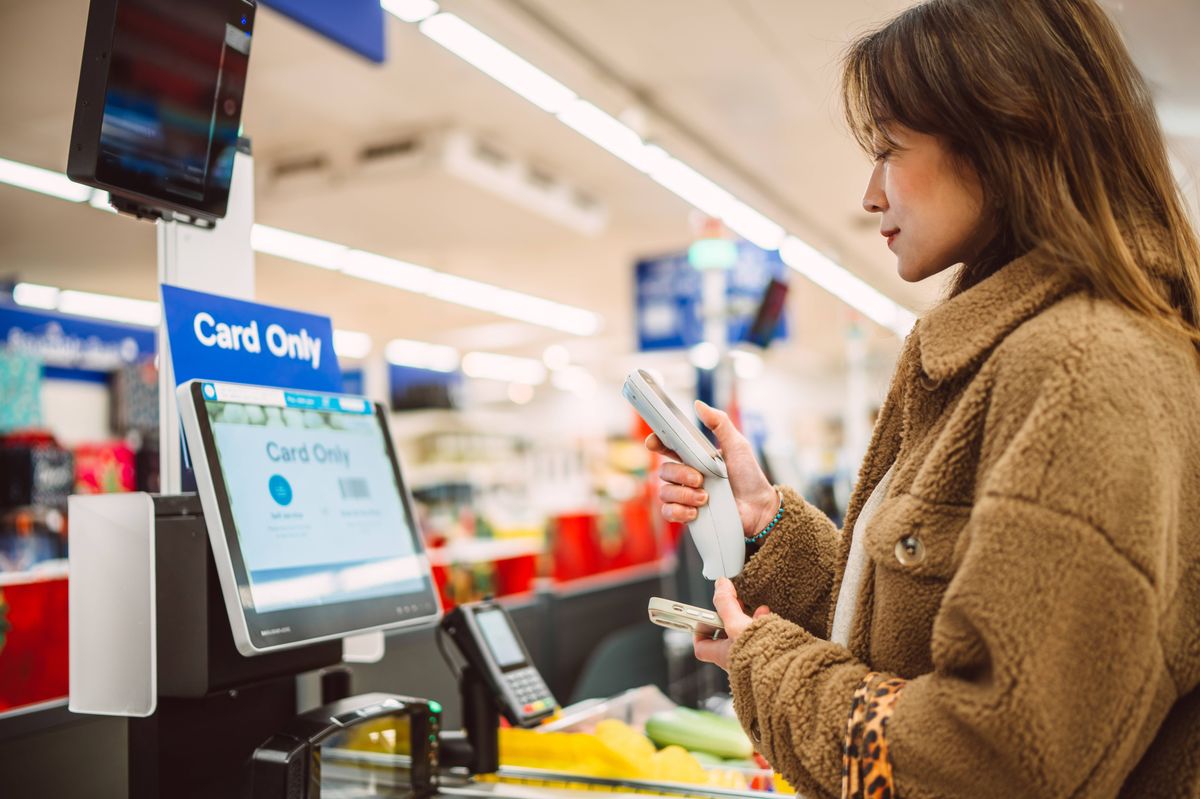 Woman using a handheld self-checkout scanner and contactless payment at a supermarket. Concepts of mobile banking, cashless shopping, e-commerce innovation, and mobile wallet.