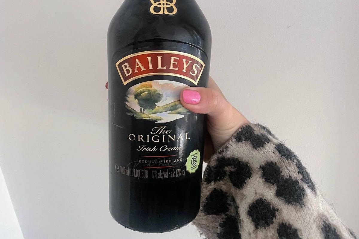 A bottle of Baileys Original Irish Cream, a dark liquor with a red label, held by a hand wearing a leopard-print glove against a plain background.