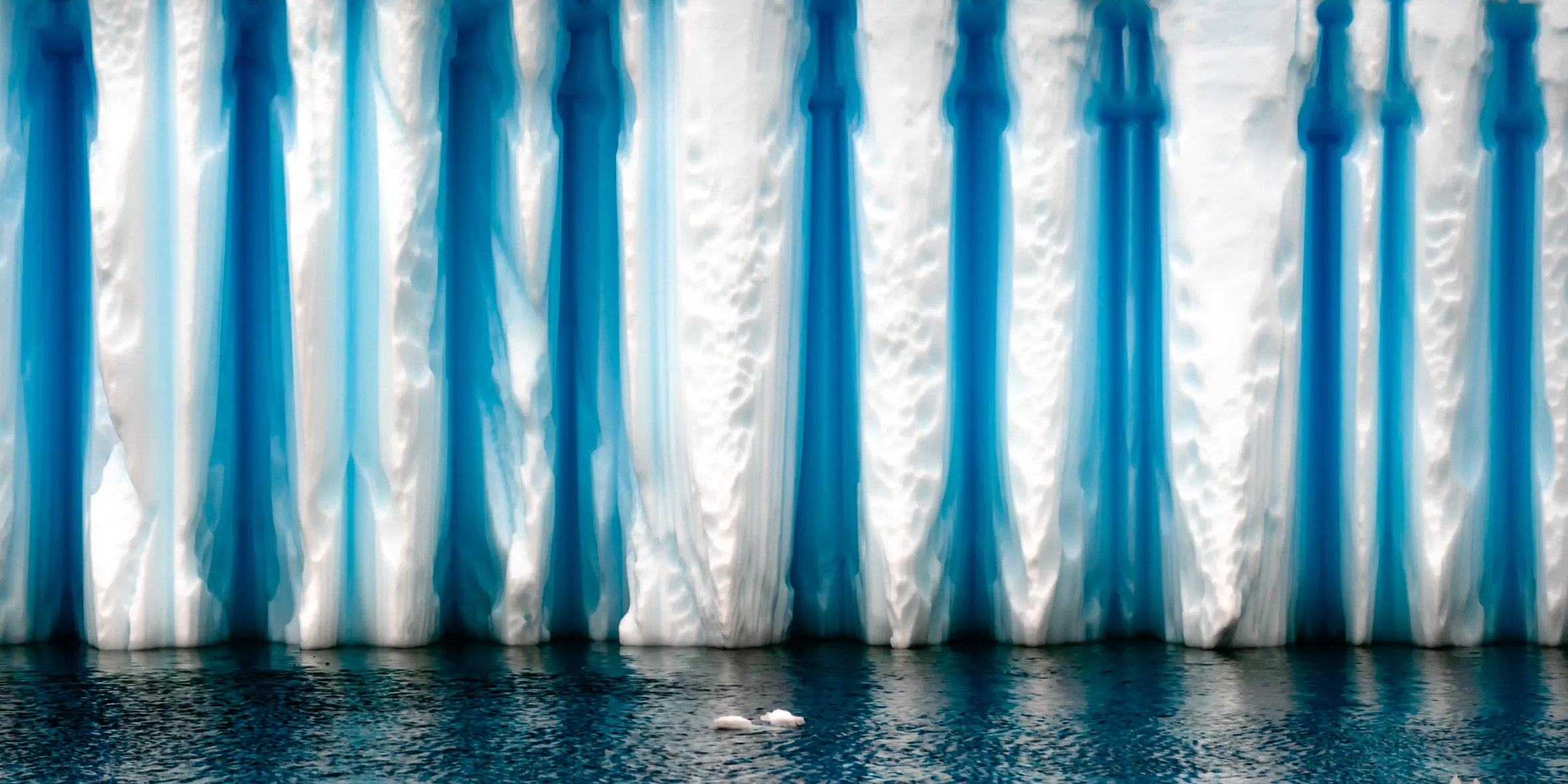 Antarctic iceberg with white and deep blue vertical stripes reflecting on the water.