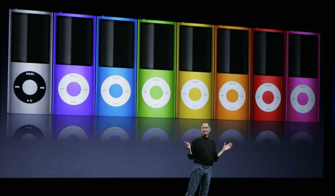 Apple's then-CEO, Steve Jobs, stands on a stage to introduce new colors for the iPod Nano in September 2008. Behind him is an enlarged image of a row of iPod Nanos in seven rainbow colors and gray.