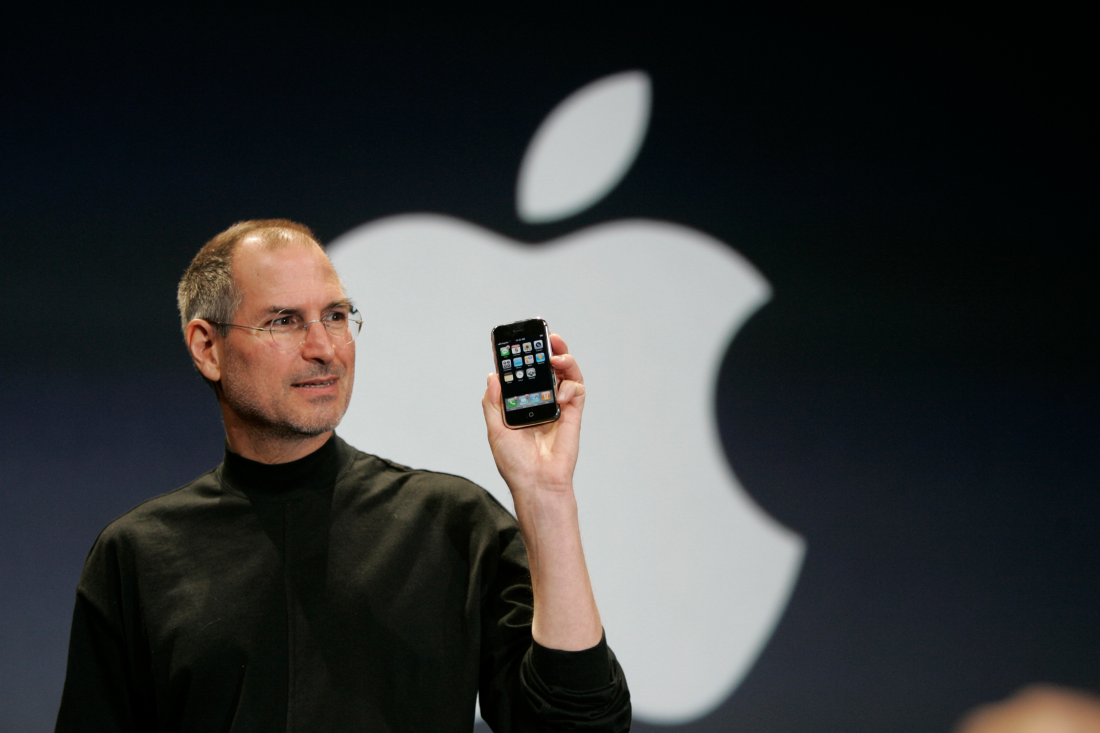In this photo, Steve Jobs, wearing his signature black turtleneck, holds up an early-model iPhone. Behind him on a black backdrop is a large white Apple logo.