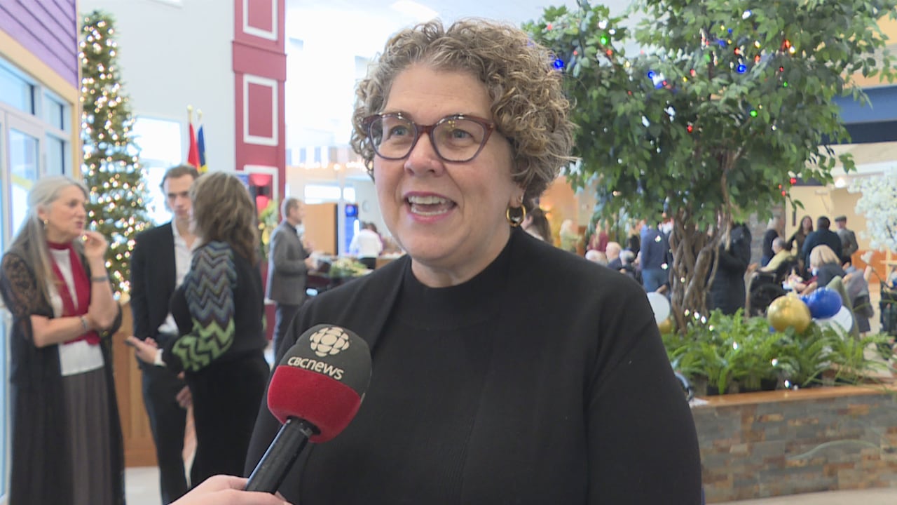A woman wearing a black turtleneck with curly short hair speaks in to a microphone at an announcement.