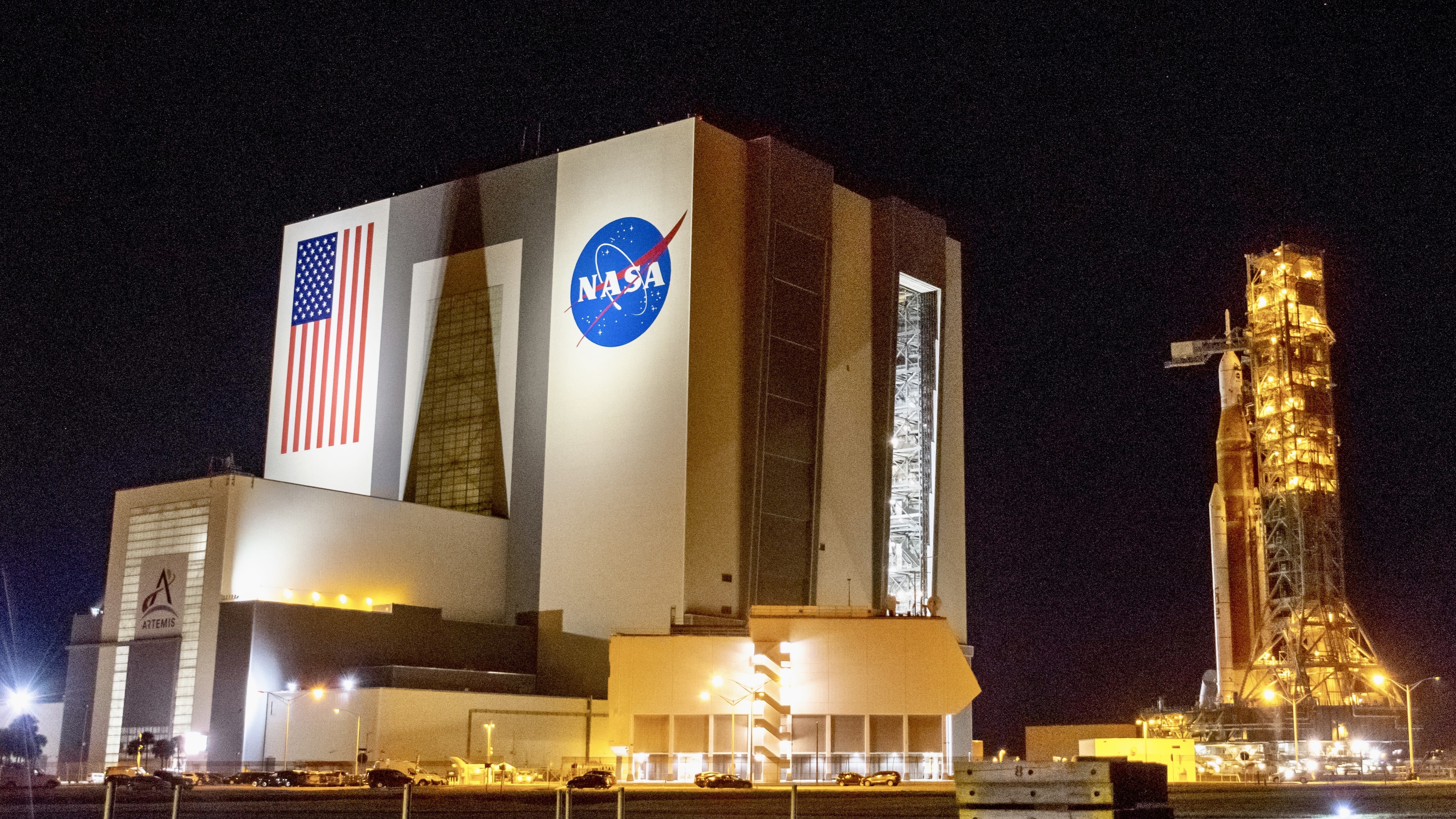 A tall orange rocket stands on a grey platform and tower next to a large cube-like building at night.