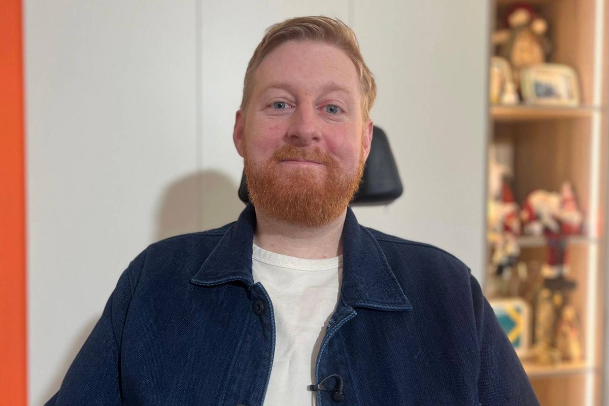 An individual with a beard is seated in a chair, dressed in a blue jacket and a white shirt, positioned in front of a white wall adorned with shelves containing various items.