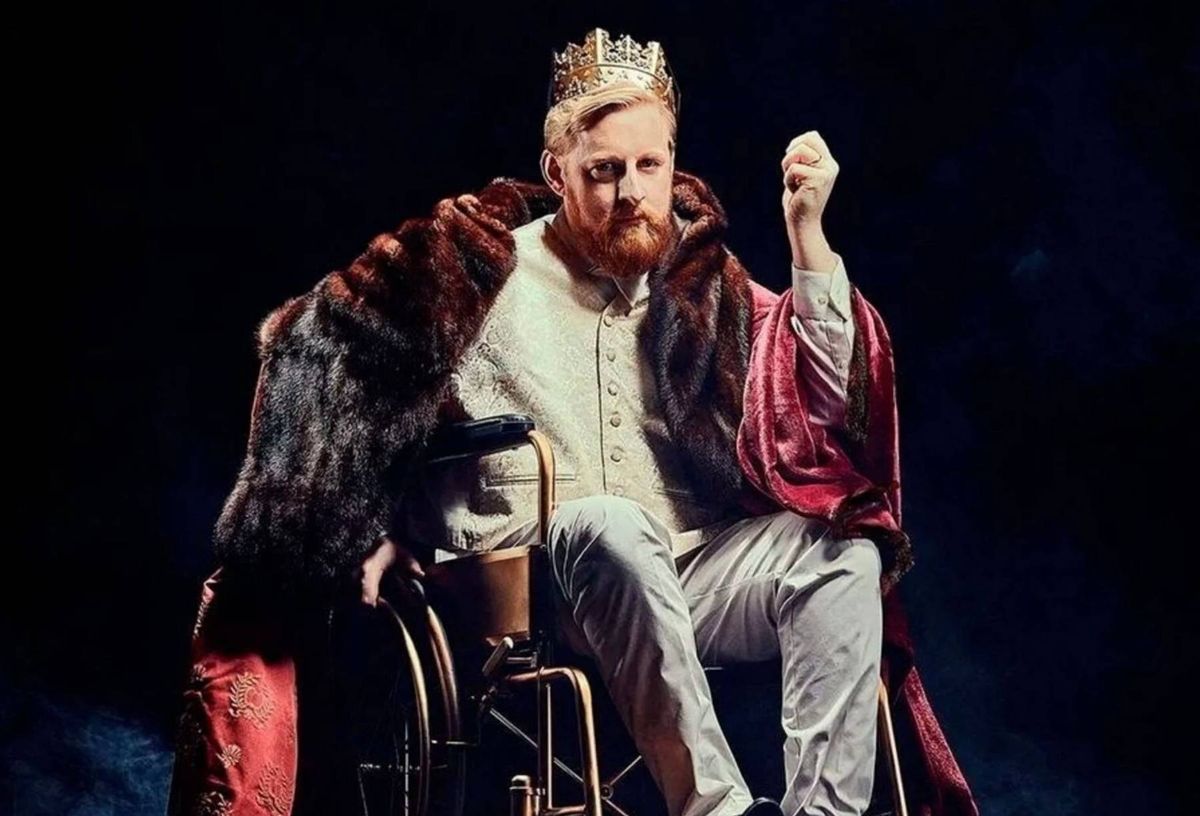 An individual wearing a crown and an ornate robe is seated in a wheelchair, raising one hand and looking forward with an expression of determination. The setting suggests a stage performance or theatrical production.