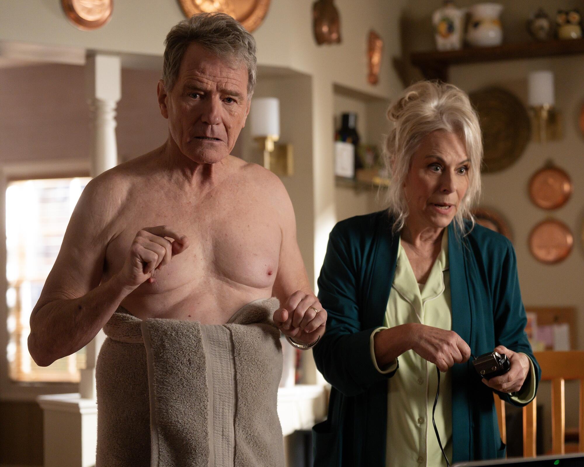 A shirtless man with a towel around his waist stands next to a woman in a green cardigan.