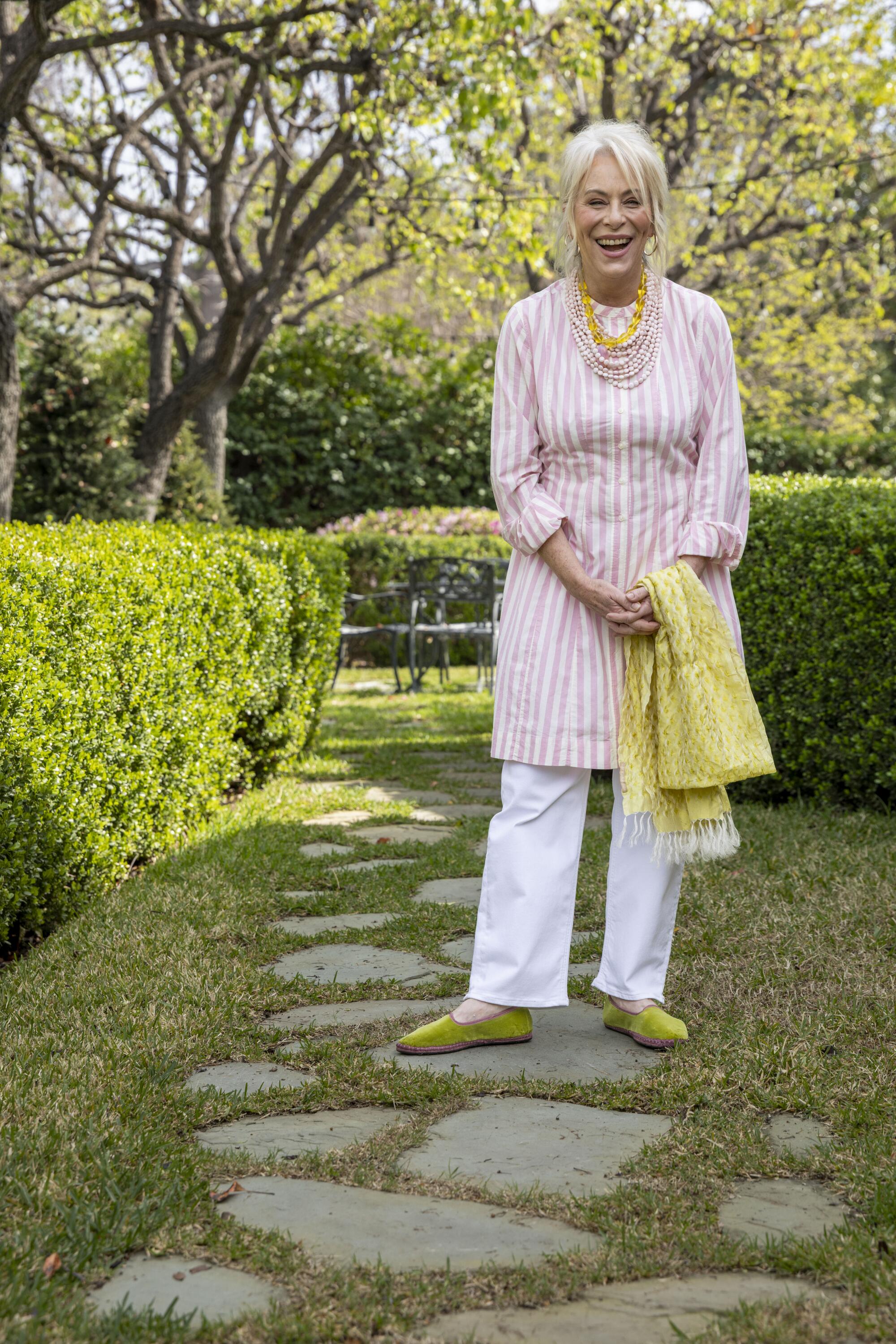 A smiling woman with white hair stands in a garden pathway.