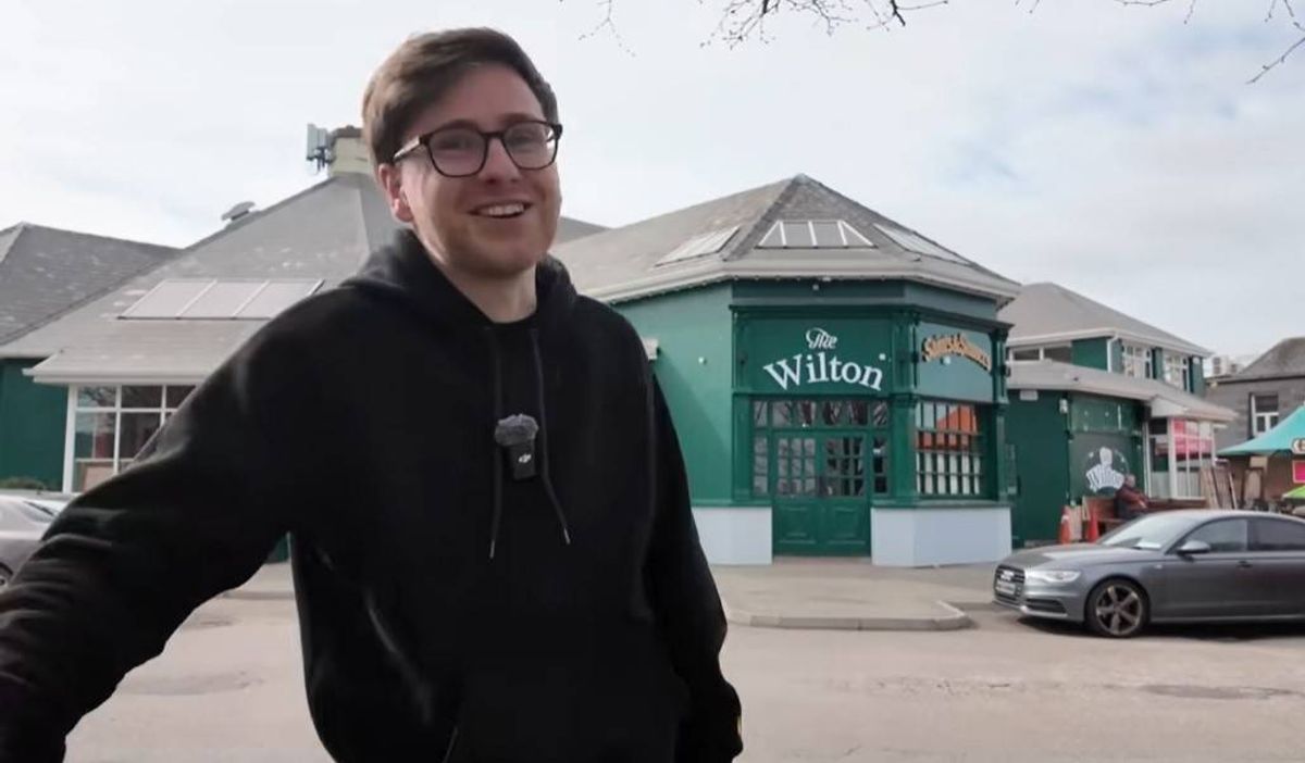 An individual dressed in a black hoodie and glasses is standing in an urban setting, with a building featuring a green facade in the background. The scene also includes parked vehicles and an urban streetscape.