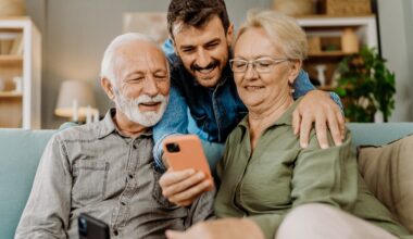 Younger Man leaning over his older parents sitting on the couch while they show pictures on a phone