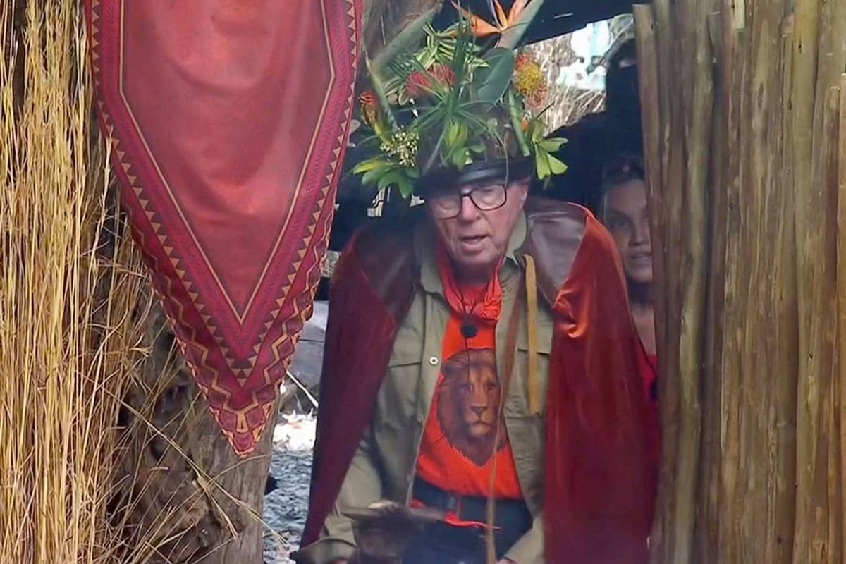 An individual adorned in a red robe and a garland of greenery is peering through a curtain embellished with red fabric.