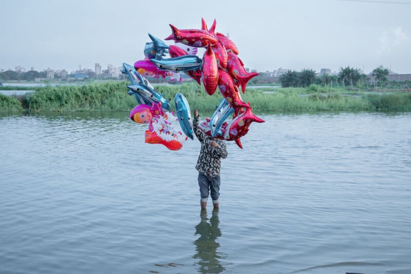 A person standing knee-deep in water holds a large bunch of colorful, cartoon-shaped balloons, obscuring their face and upper body, with greenery and distant buildings in the background.