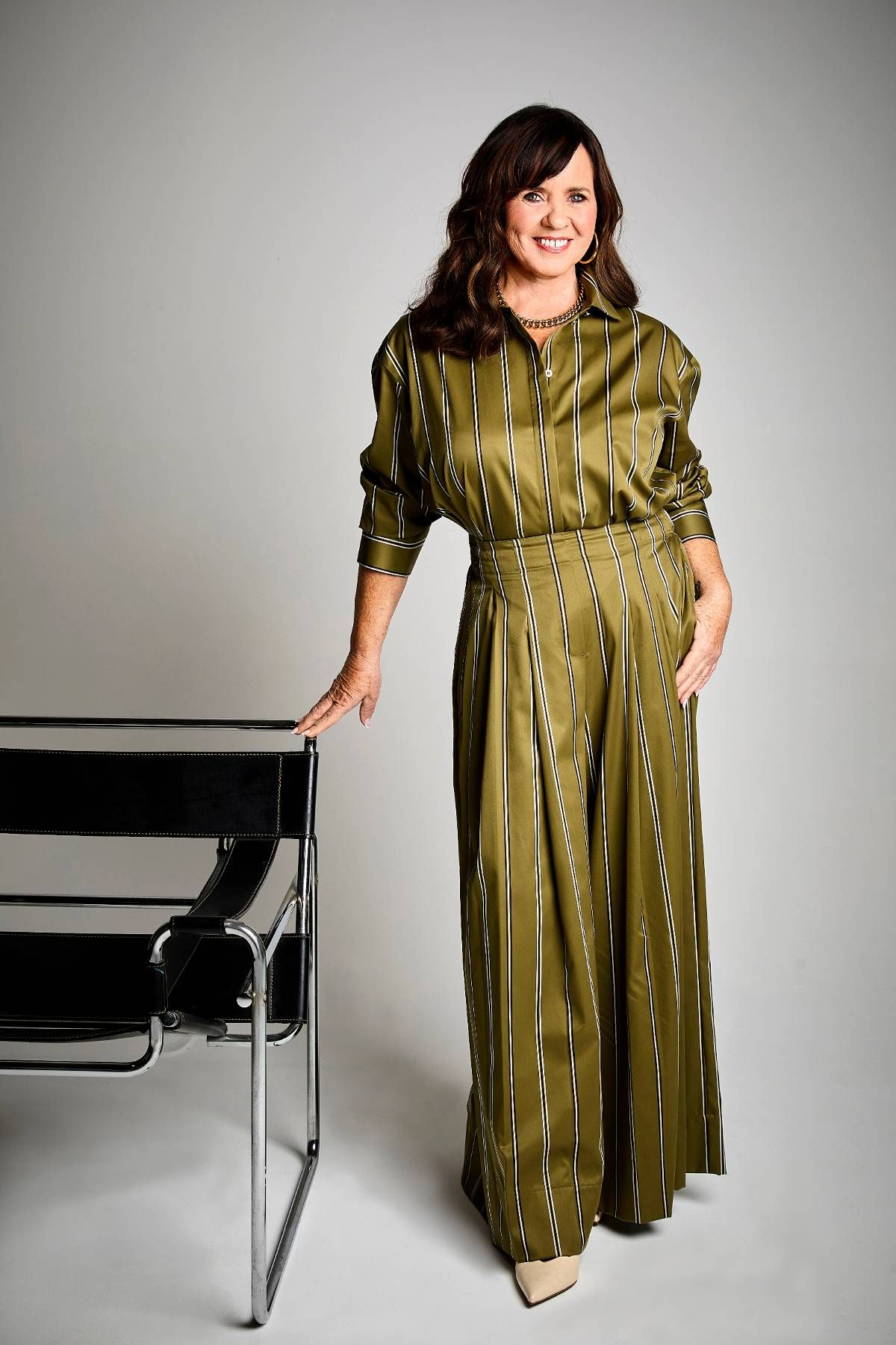 An individual is standing and smiling, dressed in a green, long-sleeved blouse with puffed sleeves against a plain gray backdrop.
