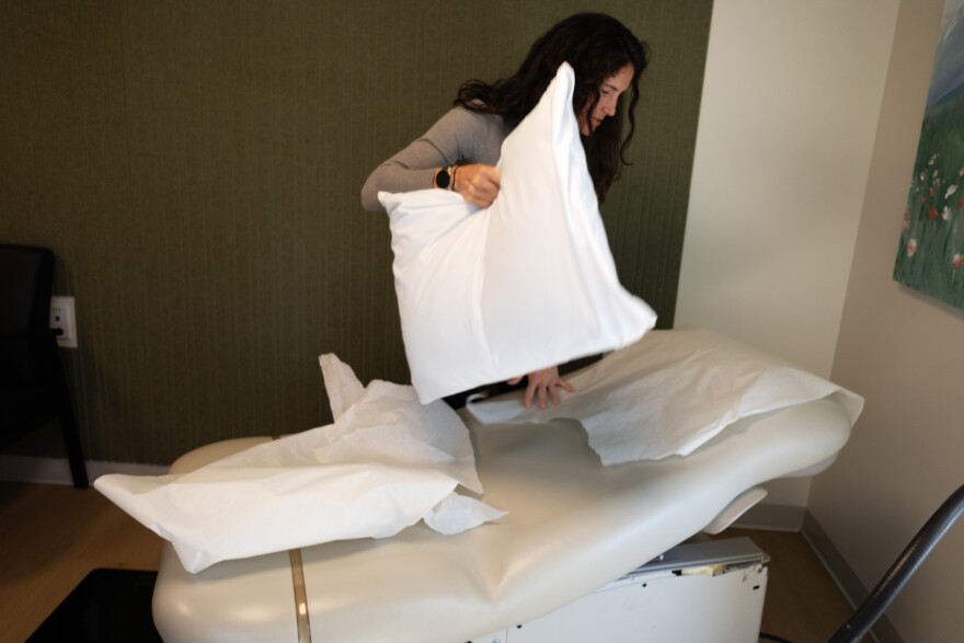 A woman with long dark hair and wearing neutral clothing changes out the exam table in a primary care exam room.