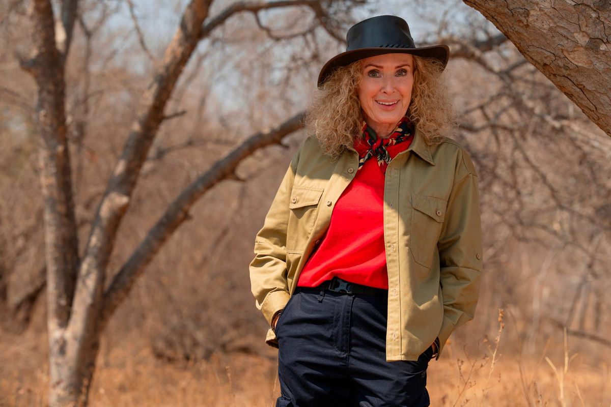 An individual dressed in a red top, black trousers, and a green and yellow jacket, with a black hat, is standing outdoors amidst a landscape of sparse trees and dry grass.