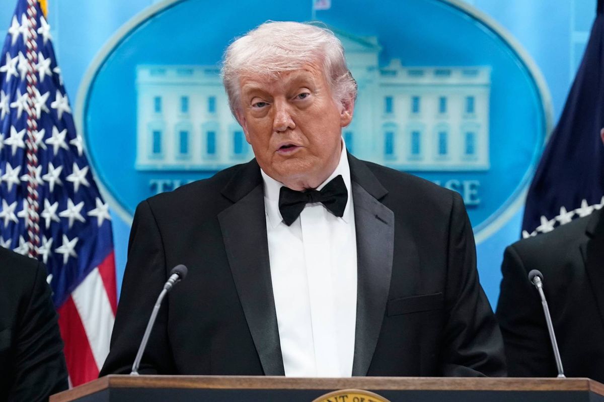 An individual dressed in formal attire, consisting of a black tuxedo and bow tie, stands at a podium, addressing an audience. Behind the speaker, the iconic backdrop of the White House is visible, with American flags positioned on either side, creating a patriotic atmosphere.