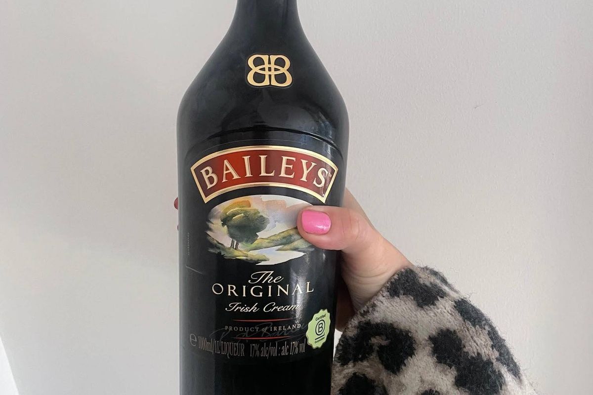 A bottle of Baileys Original Irish Cream, a dark liquor with a red label, held by a hand wearing a leopard-print glove against a plain background.