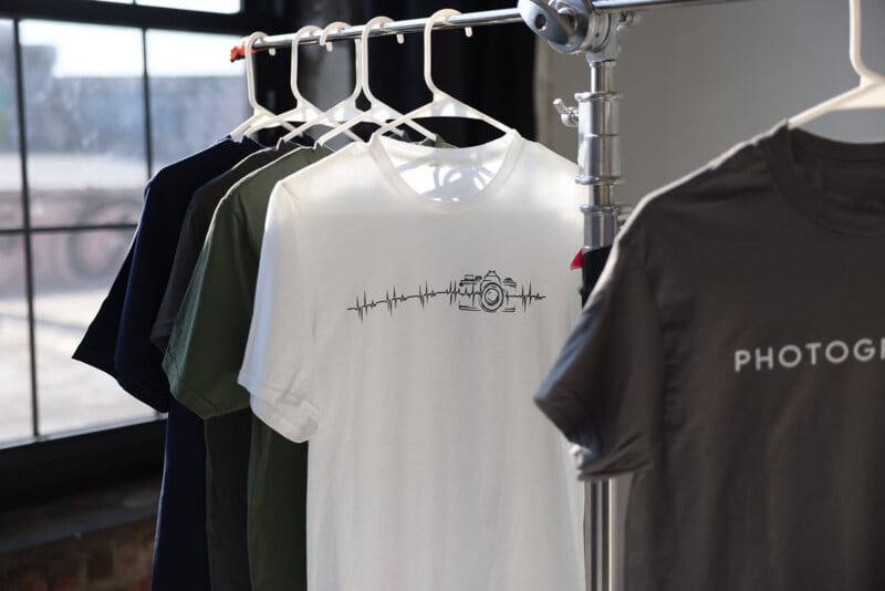 Several t-shirts hang on white hangers from a metal rack, including a white shirt with a camera and heartbeat line graphic, and a gray shirt partially visible with the word "PHOTOG" printed on it. A large window is in the background.
