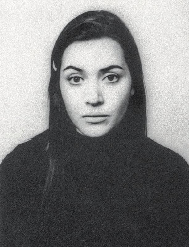 A black and white portrait of a woman with long dark hair, wearing a dark top, looking directly at the camera with a neutral expression against a plain background.