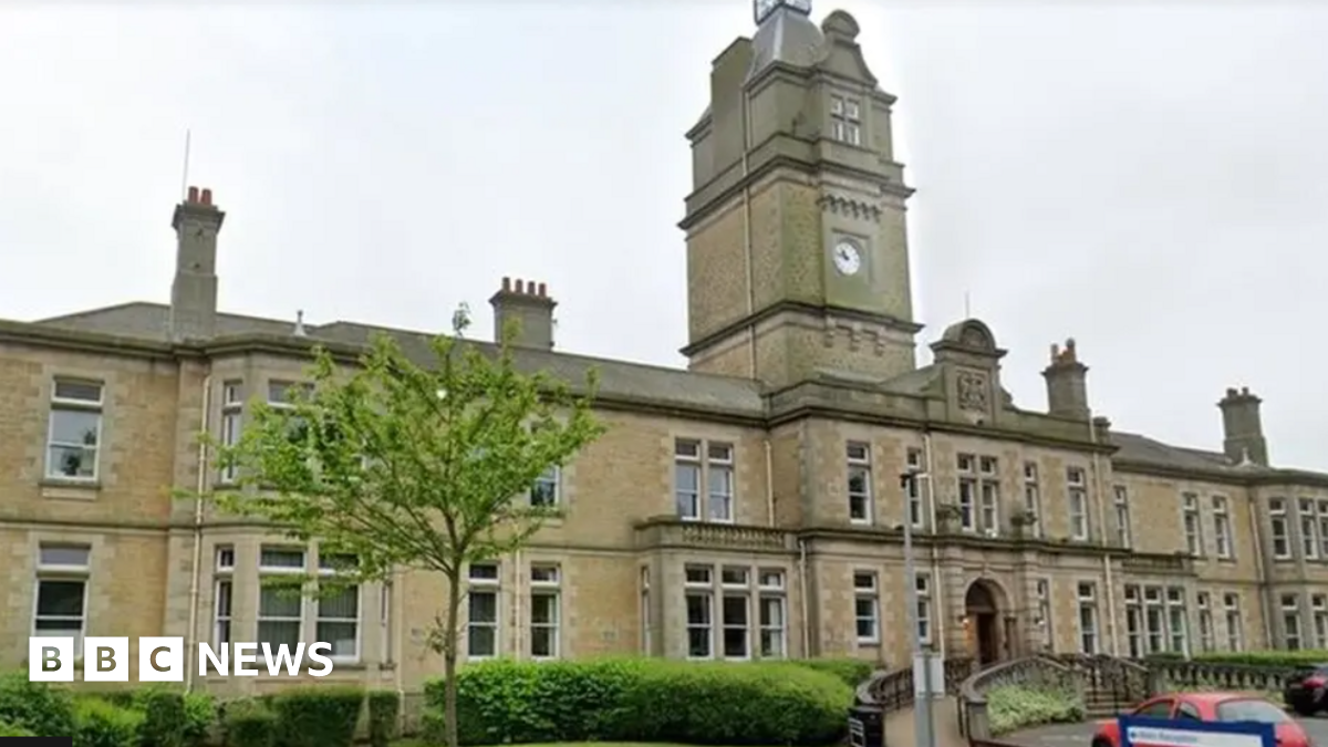 A long large Victorian building takes up most of the image with steps leading up to the entrance and a ramp to one side. A large clock tower sits in the centre of the building and there are several chimneys