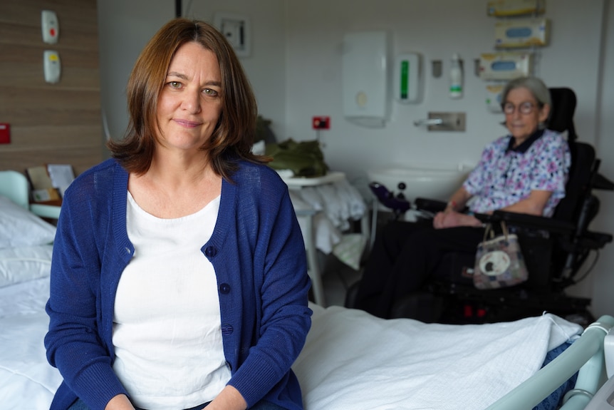 A woman with long brown hair sits on a hospital bed, a woman with grey hair and glasses in a wheelchair behind her.