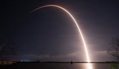 A rocket with a capsule on the end flies through the night sky, leaving a trail of white light reflecting off the water in the foreground.