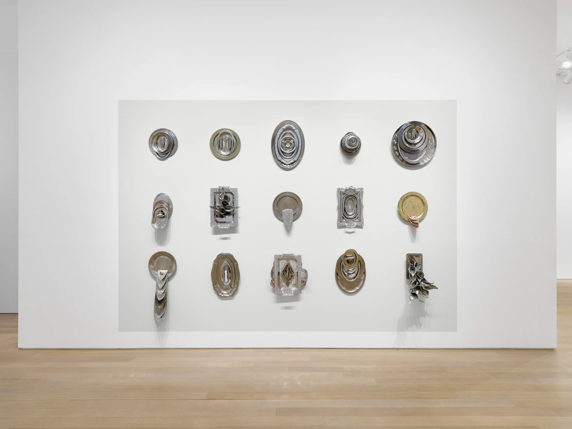 Wall-mounted abstract sculptures on a gallery wall displayed in a grid pattern with various shapes and textures.