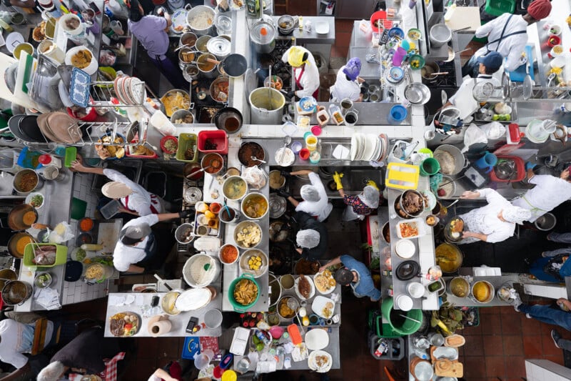 A busy overhead view of chefs working in separate kitchen stations, filled with pots, pans, plates, and ingredients, creating a vibrant and chaotic cooking environment.