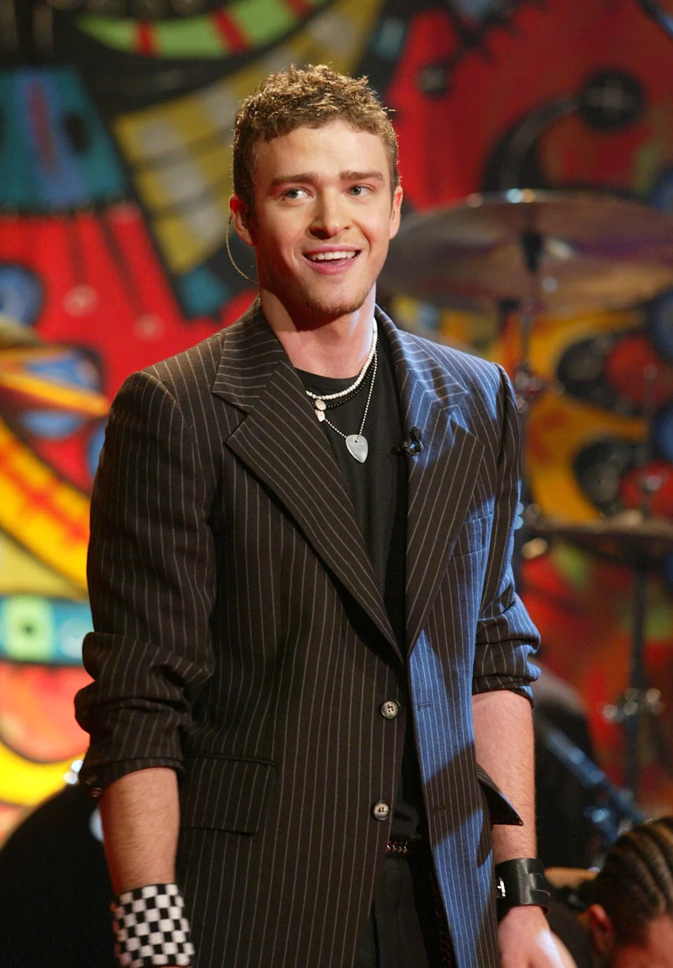 A person is standing with short curly hair, wearing a pinstripe jacket, black shirt, layered necklaces, and checkered wristbands, against a colorful backdrop