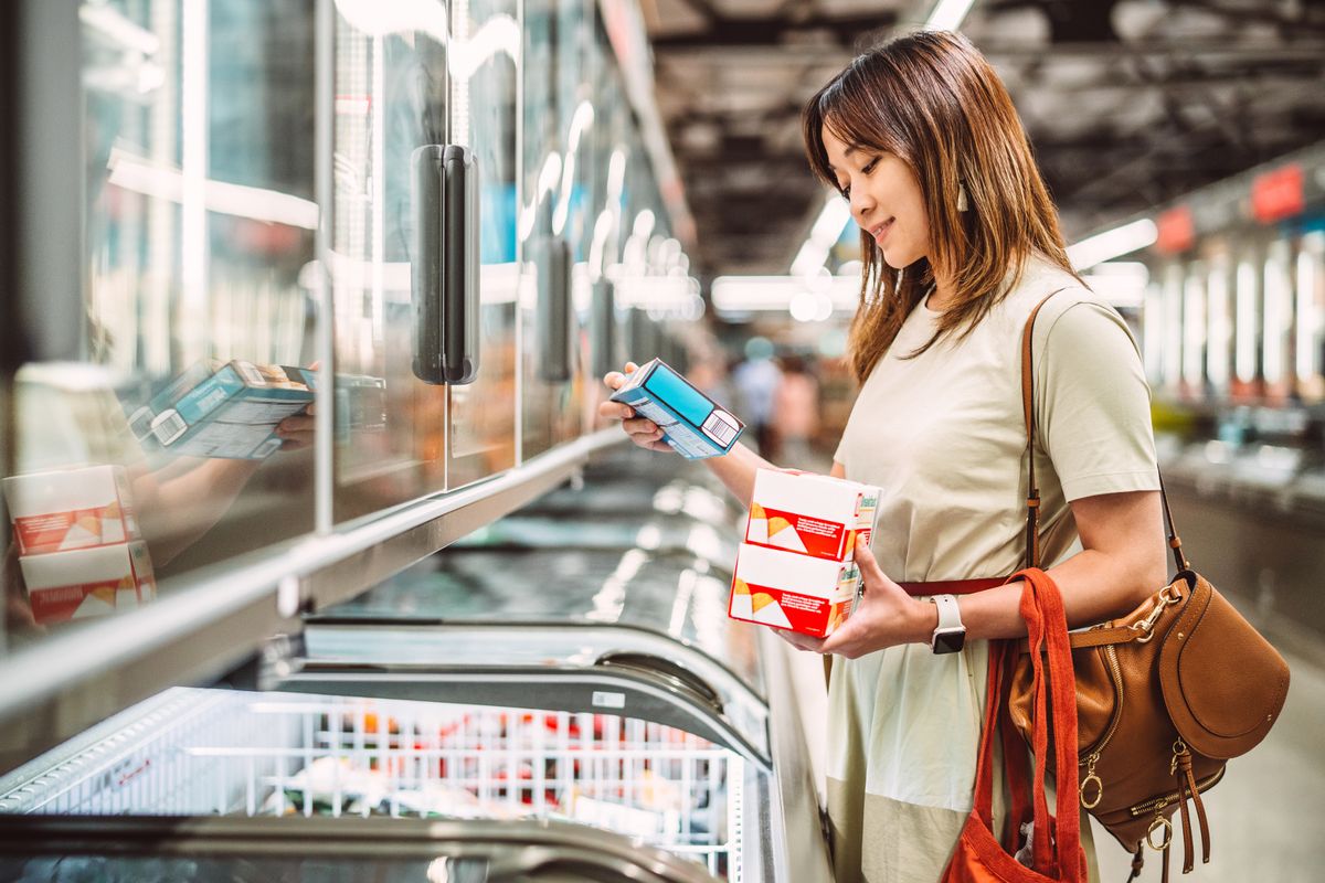 Woman selecting packaged food from a supermarket freezer aisle while grocery shopping. Concept of consumer choice, cost of living, food shopping, and nutrition decision.