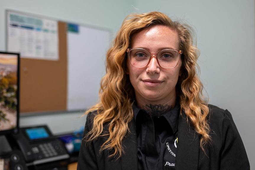 A woman with red hair and glasses smiling, with a desk and corkboard behind her