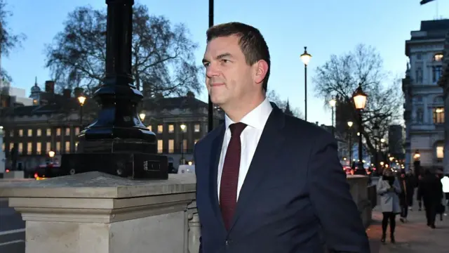Olly Robbins walking along a road in front of street lights as it's dusk. He smiles with his mouth closed