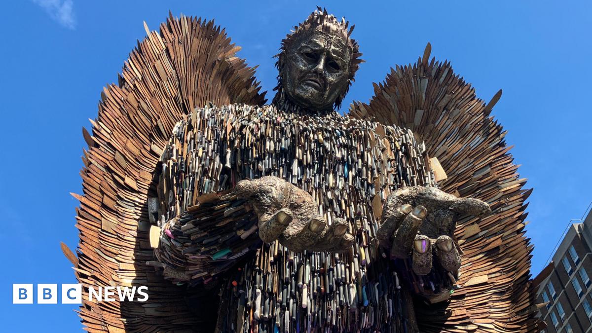 A tall male figure towers above a bright blue sky. The statues hands are open and shoulders are shrugged. The statue is made out of rusty knife blades of all different shapes and sizes.