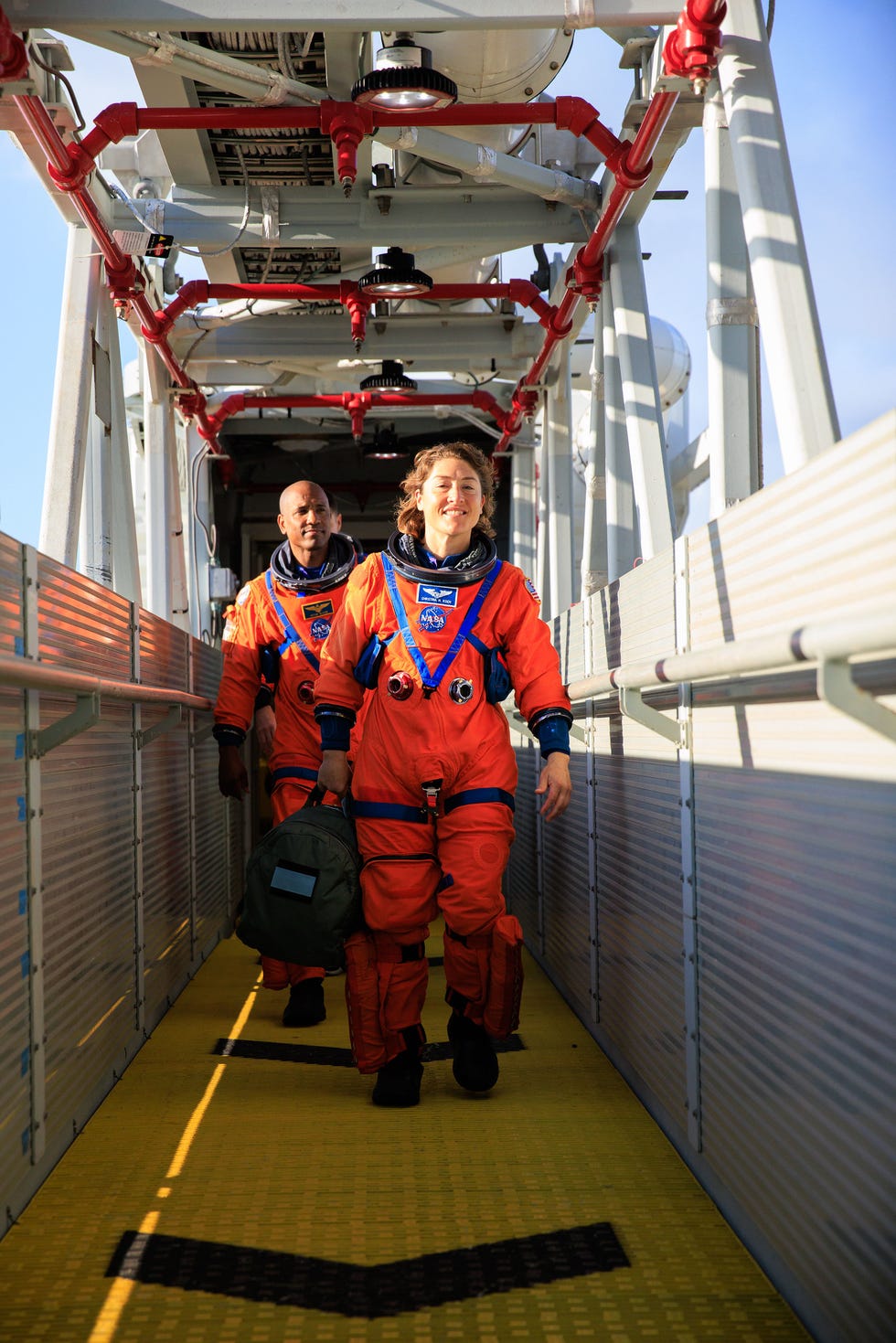 Two astronauts in orange suits walking on a bridge towards a spacecraft.
